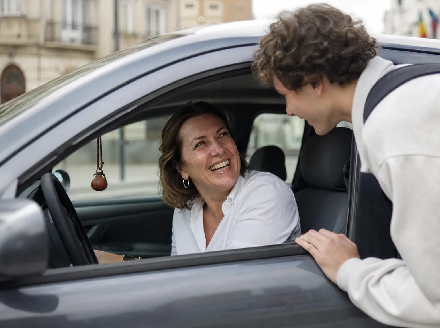 Vue par la fenêtre ouverte, une conductrice de taxi rit avec un jeune passager qui monte à bord, sac au dos.