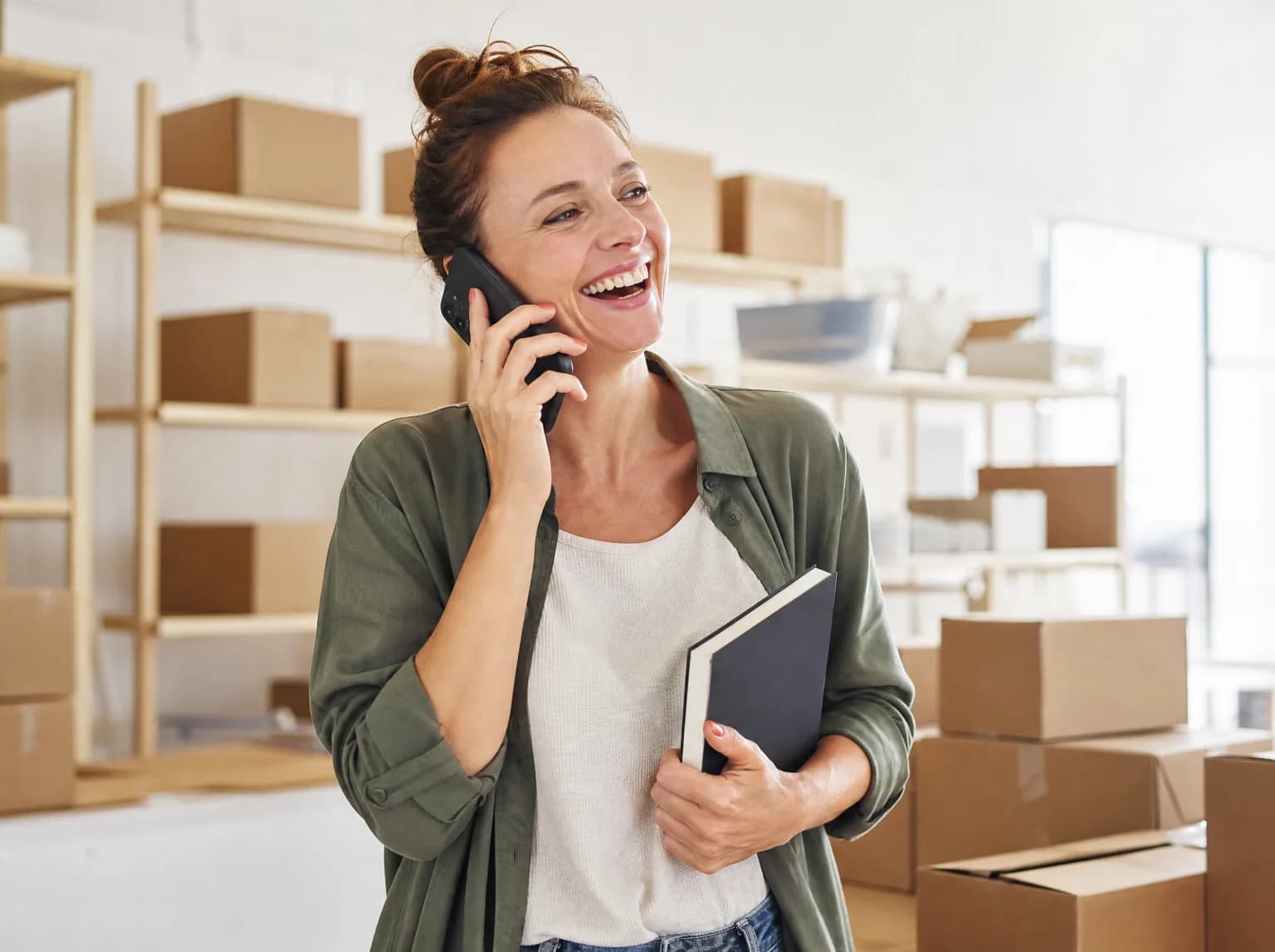 Une repreneuse rit au téléphone dans l'entrepôt lumineux de son entreprise, carnet à la main.