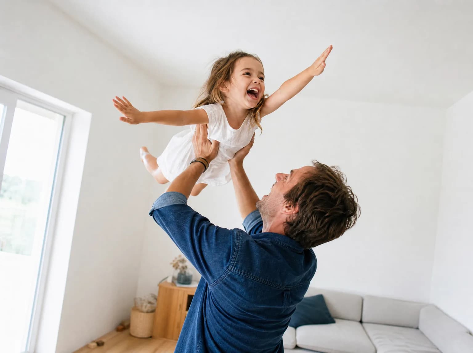 Un père soulève sa fille en riant dans un salon lumineux, les bras de l'enfant grands ouverts.