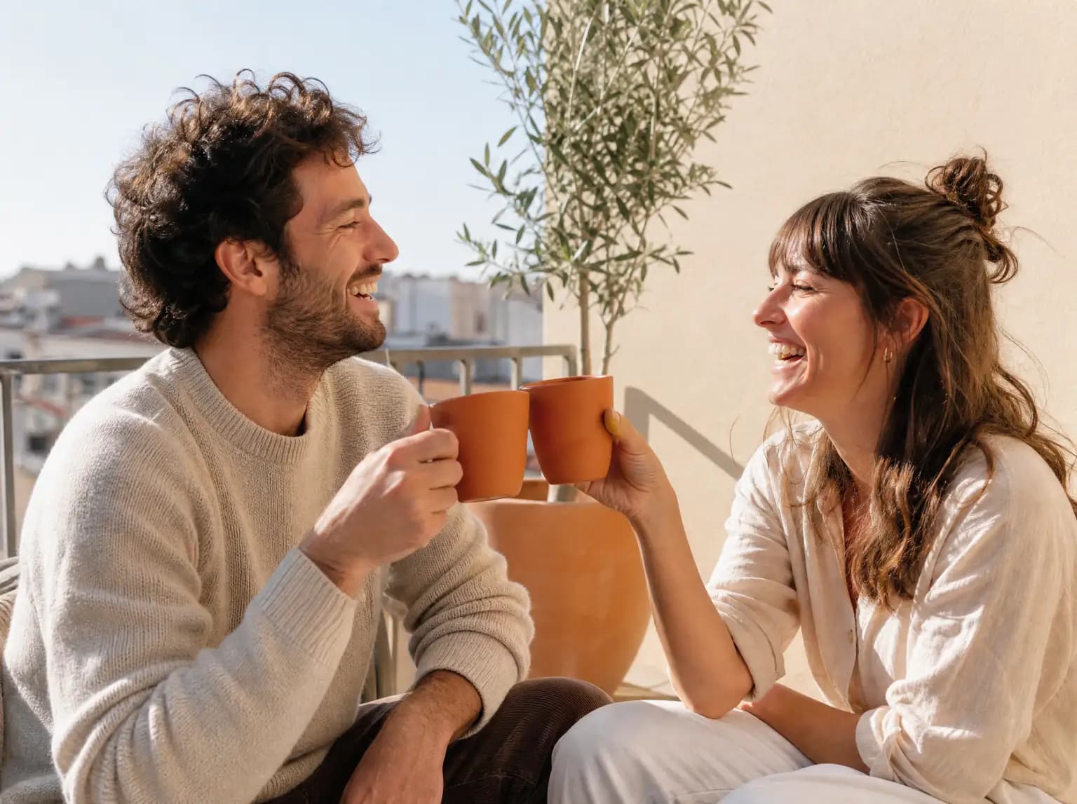 Un couple d'une trentaine d'années trinque avec deux tasses sur un balcon ensoleillé.